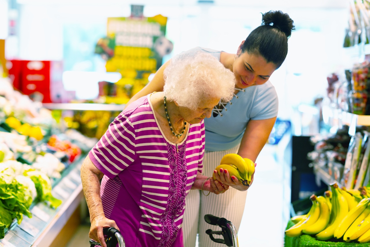 Caregiver supporting senior adult woman with buying bananas in a shop.