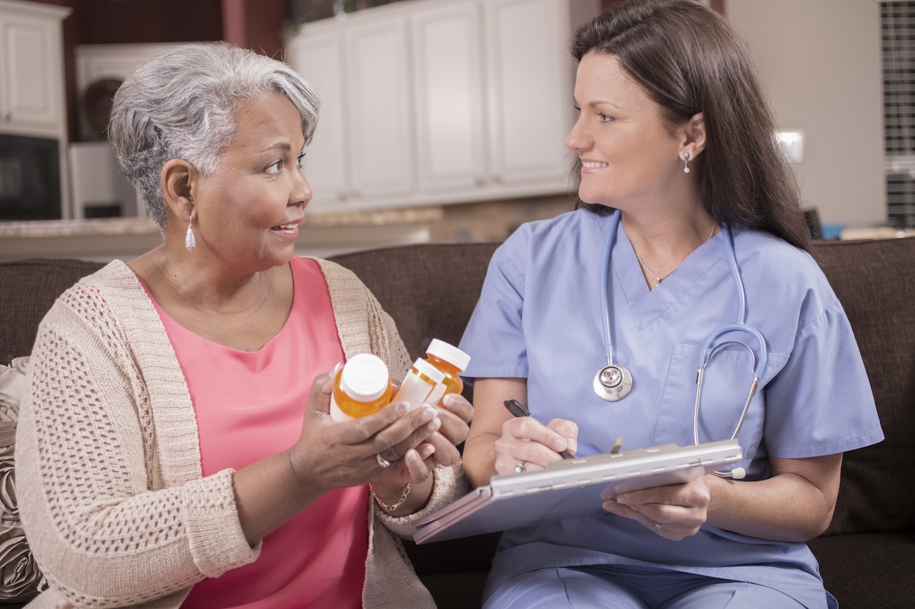 Caring healthcare nurse discusses prescription medications with senior adult patient at home, assisted living, or nursing home setting. She holds a clipboard as she explains the medication information. Kitchen background.
