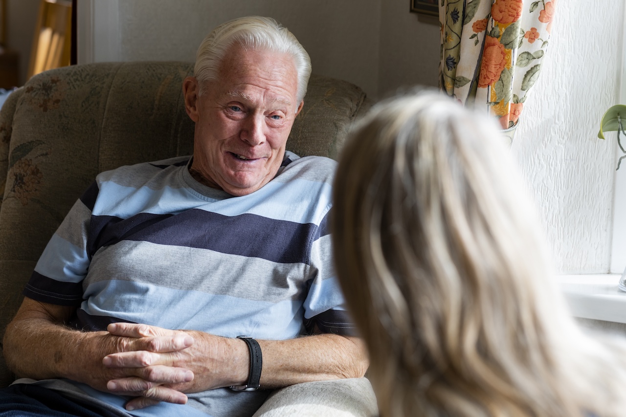 Nurse conversing with a senior adult patient while he is sitting in a chair in his living room.