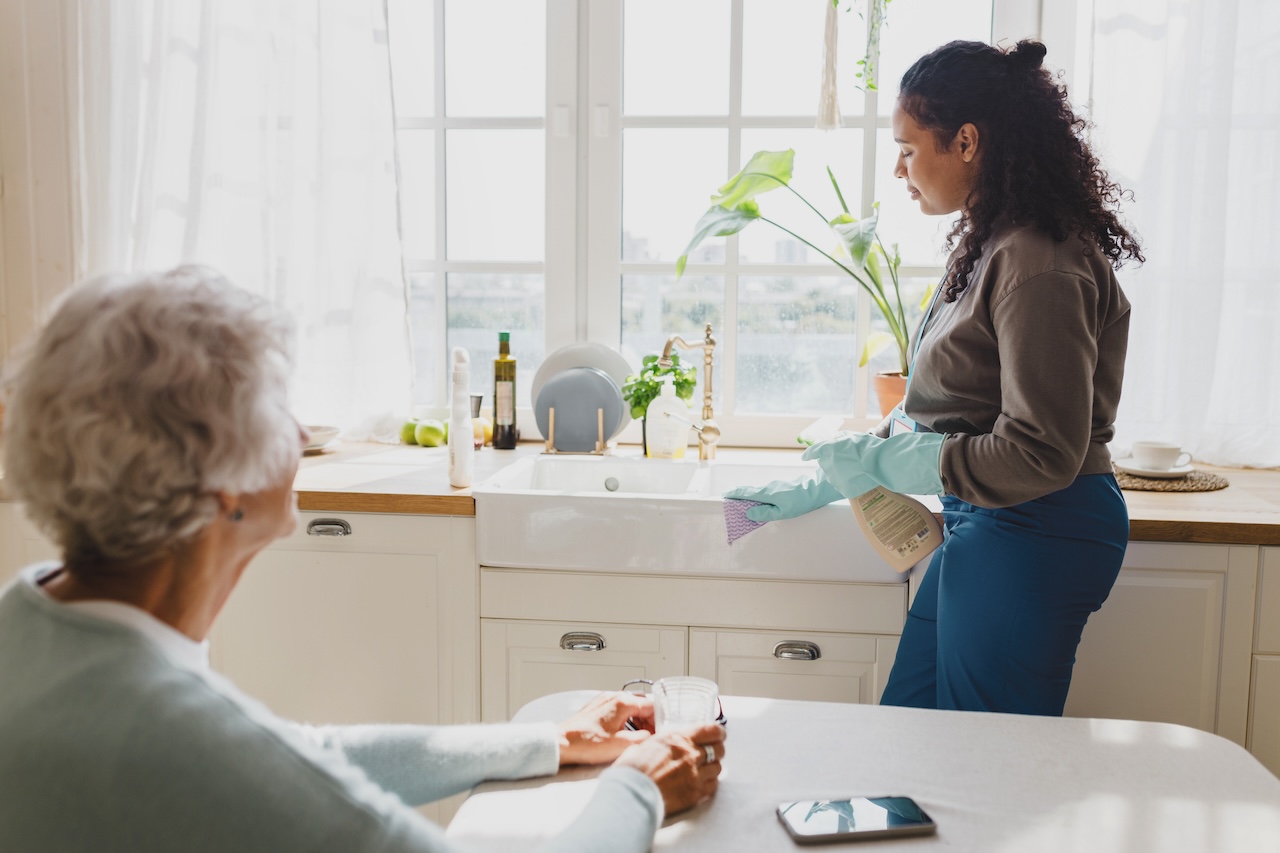 Senior adult lady with gray hair looking at housekeeping volunteer helping to clean house, washing kitchen sink with detergent, wearing gloves, talking to each other