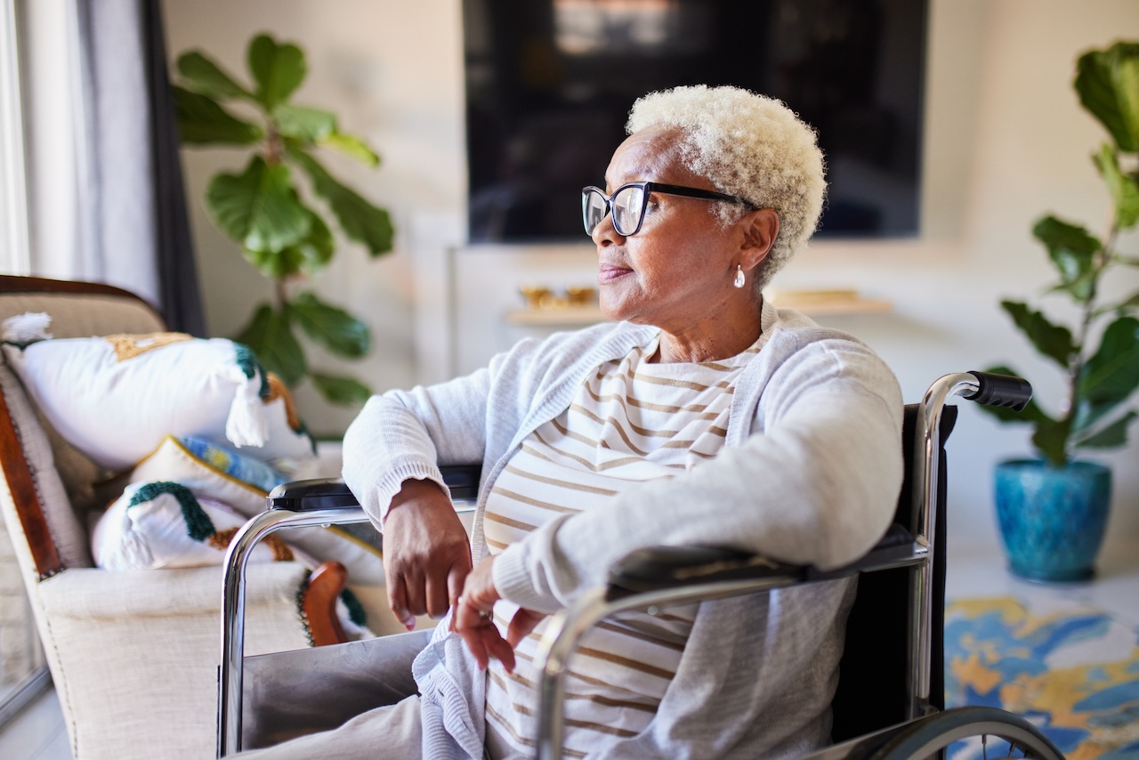 Senior adult woman with white hair and glasses sits in a wheelchair, looking out the window. She wears a striped shirt and a cardigan. Potted plants and a patterned rug are visible in the background.