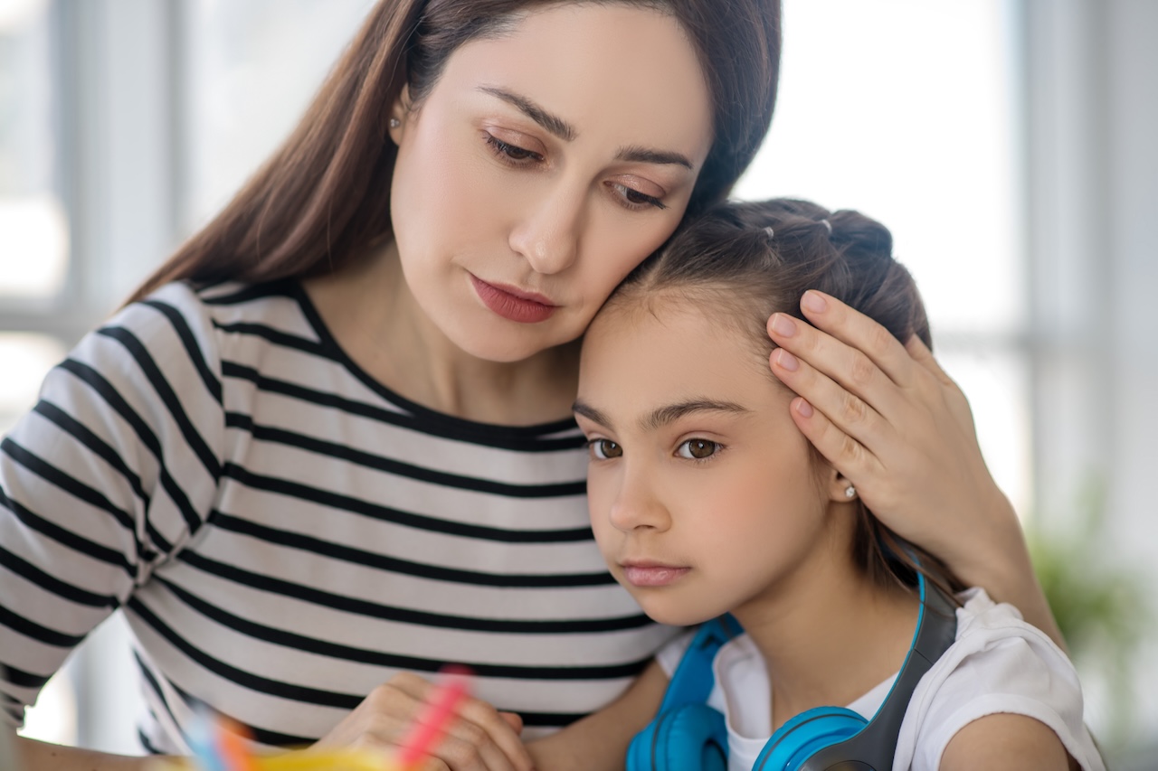 Young loving caring mother hugging her sad daughter, both sitting at home at the table.